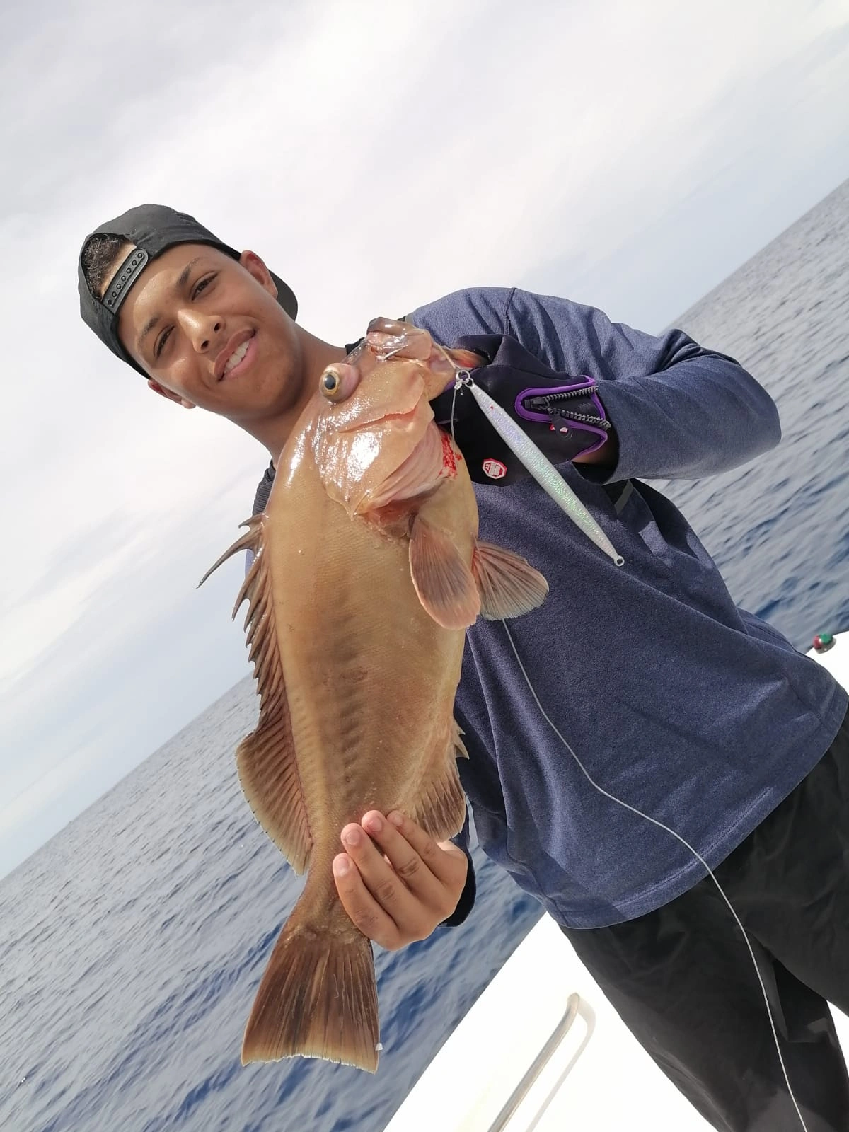 Captura de Mero - En las profundidades de Coiba, un pescador experto ha logrado capturar un imponente mero utilizando un jig de 80 gramos. La fuerza y habilidad del pescador se reflejan en la captura, mientras el mero brilla con sus intensos colores, símbolo de la riqueza marina de la región.
